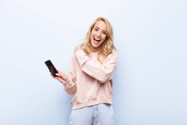 Genç sarışın kadın mutlu, pozitif ve başarılı hissediyor, meydan okumayla karşılaştığında motive oluyor veya akıllı telefon kullanarak iyi sonuçları kutluyor.