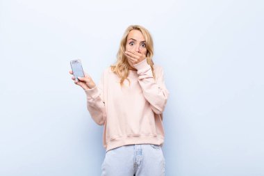 Şok olmuş, şaşırmış bir ifadeyle ağzı kapalı genç sarışın kadın bir sır saklıyor ya da akıllı telefon kullanıyor.