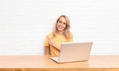 Genç sarışın kadın neşeyle gülümsüyor, mutlu hissediyor ve yan ve yukarı doğru işaret ediyor, fotokopi uzayında dizüstü bilgisayar kullanarak nesneleri gösteriyor.