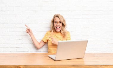 Genç sarışın kadın şaşkın ve şaşırmış hissediyor, şok olmuş bir ifadeyle gülümsüyor ve dizüstü bilgisayar kullanarak yüzünü işaret ediyor.