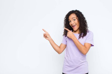 Genç siyah kadın mutlu ve şaşırmış hissediyor, şok olmuş bir ifadeyle gülümsüyor ve beyaz duvara doğru işaret ediyor.