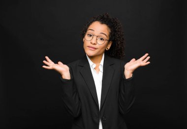 Genç siyah kadın şaşkın ve şaşkın hissediyor, şüpheci, farklı seçenekleri tartıyor veya seçiyor siyah duvara karşı komik bir ifadeyle