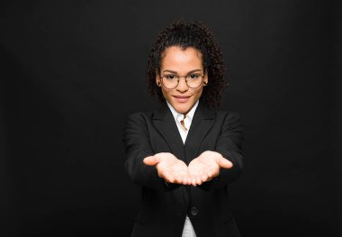 Genç siyah kadın mutlu bir şekilde gülümsüyor. Dost canlısı, kendinden emin, pozitif bakışlı, siyah duvara karşı bir nesne ya da konsept sunuyor ve gösteriyor.