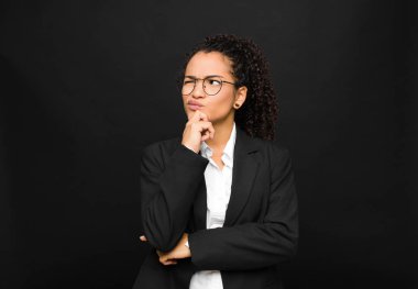 Genç siyahi kadın düşünüyor, kuşkulu ve şaşkın hissediyor, farklı seçeneklerle, siyah duvara karşı hangi kararı vereceğini merak ediyor.