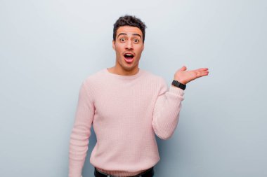 Genç Arap adam şaşırmış ve şoka uğramış görünüyor, çenesi düşmüş, gri duvara karşı eli açık bir nesneyi tutuyor.