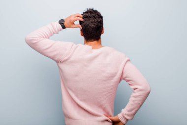 Genç Arap adam bilgisiz ve şaşkın hissediyor, bir çözüm düşünüyor, eli kalçasında, diğeri kafasında, gri duvara karşı dikiz görüşlü.