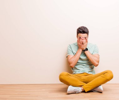 Genç Arap bir adam üzgün, hayal kırıklığına uğramış, gergin ve depresif, iki eliyle yüzünü kapatmış, yerde oturmuş ağlıyor.