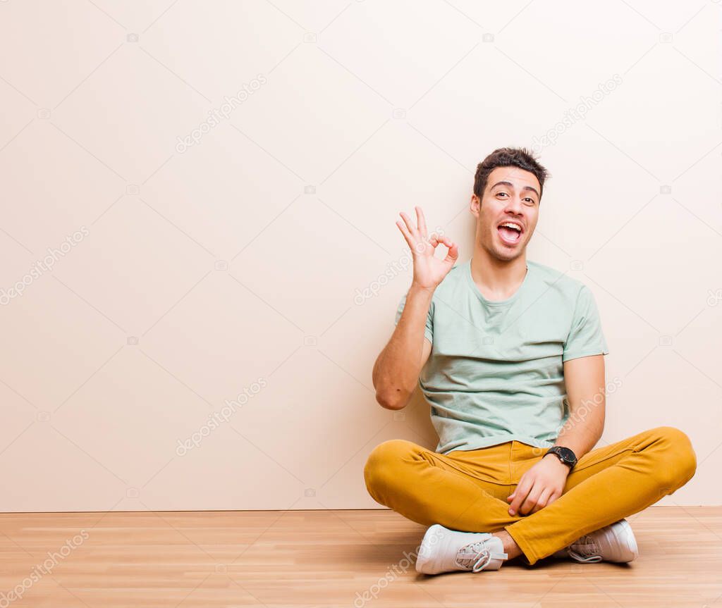 young arabian man feeling successful and satisfied, smiling with mouth wide open, making okay sign with hand sitting on the floor