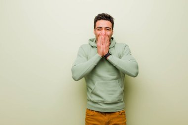 Genç Arap adam mutlu, neşeli, şanslı ve şaşırmış görünüyor. İki eli de düz duvarda.