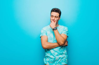 Genç Arap bir adam mutlu, kendinden emin bir ifadeyle gülümsüyor, elleri çenesinde, merak ediyor ve düz duvara bakıyor.