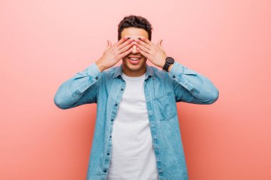 Genç Arap adam gülümsüyor ve mutlu hissediyor, iki eliyle gözlerini kapatıyor ve pembe duvara karşı inanılmaz bir sürpriz bekliyor.