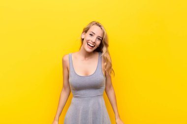 Genç, sarışın bir kadın, büyük, dost canlısı, kaygısız bir gülümseme, pozitif görünüyor, rahat ve mutlu, turuncu duvarda takılıyor.