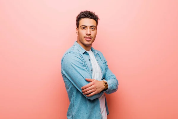 young arabian man smiling to camera with crossed arms and a happy, confident, satisfied expression, lateral view against pink wall