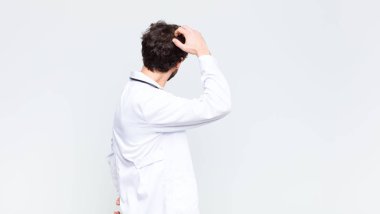 Genç doktor düşünüyor ya da şüphe ediyor, kafasını kaşıyor, kafası karışık ve kafası karışık hissediyor, fotokopi duvarının arkasında ya da arkasında.