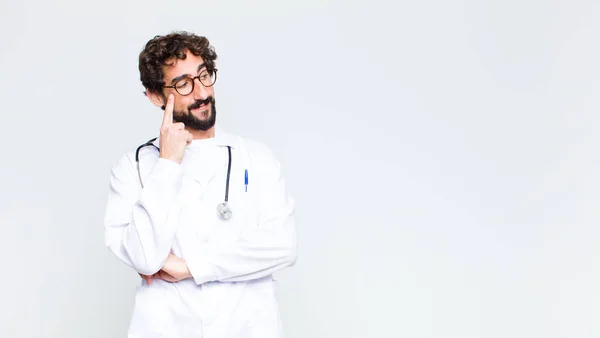 young doctor man with a concentrated look, wondering with a doubtful expression, looking up and to the side against copy space wall