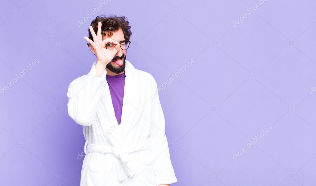 young bearded man wearing bathrobe smiling happily with funny face, joking and looking through peephole, spying on secrets