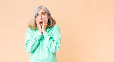 Orta yaşlı havalı bir kadın şok olmuş ve korkmuş hissediyor, ağzı açık ve elleri yanaklarında korkmuş görünüyor.
