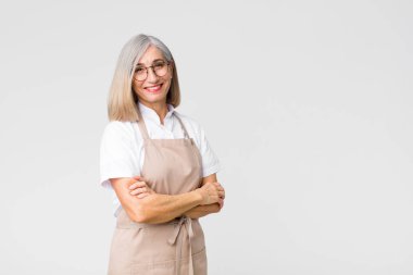 Orta yaşlı fırıncı kadın kameraya çapraz kollarıyla gülümsüyor ve mutlu, kendinden emin, tatmin olmuş bir yüz ifadesi düz duvara karşı yanal bir bakış.