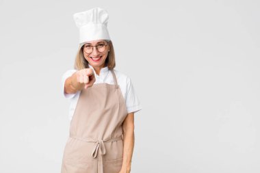 Orta yaşlı fırıncı kadın kameraya tatmin olmuş, kendine güvenen, dost canlısı bir gülümsemeyle bakıyor, düz duvara karşı seni seçiyor.