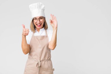 Orta yaşlı fırıncı kadın kendini mutlu, şaşkın, tatmin olmuş ve şaşırmış hissediyor, iyi ve güzel hareketler gösteriyor, düz duvara gülümsüyor.