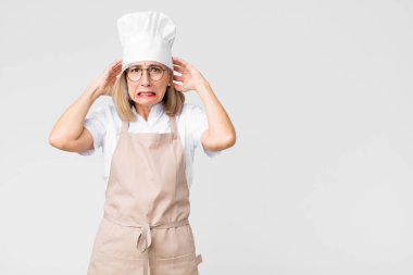 Orta yaşlı fırıncı kadın kendini stresli, endişeli ya da korkmuş hissediyor, elleri başının üstünde, düz duvara karşı panikliyor.