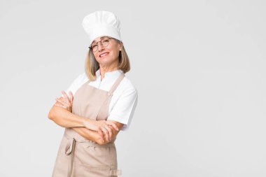 Orta yaşlı fırıncı kadın kollarını kavuşturup mutlu bir şekilde gülüyor, rahat, pozitif ve tatmin olmuş bir şekilde düz duvara dayanıyor.