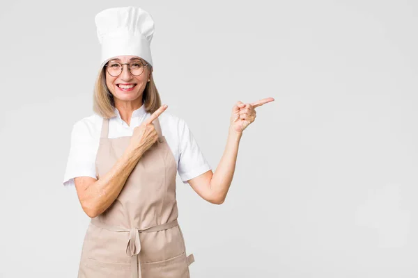 中年のパン屋の女性は笑顔で横と上を指します両手でオブジェクトを示します平らな壁に対してコピースペース — ストック写真