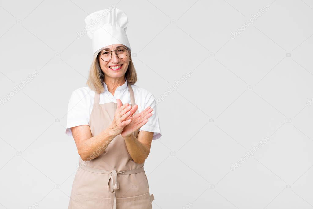 middle age baker woman feeling happy and successful, smiling and clapping hands, saying congratulations with an applause against flat wall