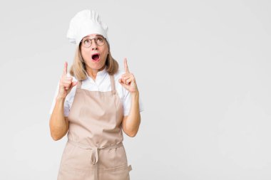 Orta yaşlı fırıncı kadın dehşete kapılmış ve ağzı açık bir şekilde yukarı doğru bakıyor. Şok olmuş ve şaşırmış bir şekilde düz duvara bakıyor.