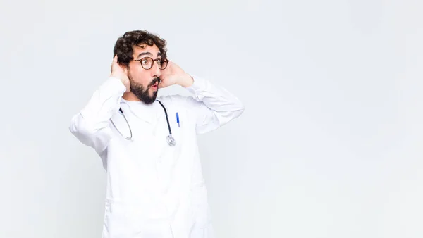 young doctor man with open mouth, looking horrified and shocked because of a terrible mistake, raising hands to head against copy space wall