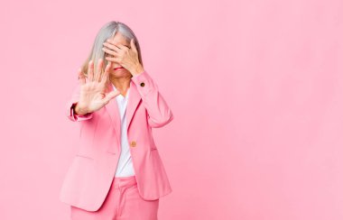 Orta yaşlı, havalı bir kadın yüzünü elle kapatıyor ve diğer elini kamerayı durdurmak için öne koyuyor. Duvara karşı fotoğraf ya da resim çekmeyi reddediyor.