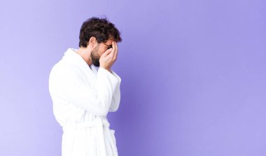 Bornoz giymiş genç sakallı bir adam üzgün, hayal kırıklığına uğramış bir bakışla gözlerini kapatıyor, ağlıyor, yan gözle bakıyor.