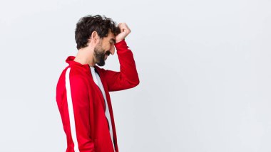 young bearded man back view raising palm to forehead thinking oops, after making a stupid mistake or remembering, feeling dumb against copy space wall