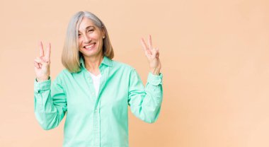 Orta yaşlı havalı bir kadın gülümsüyor ve mutlu görünüyor, arkadaş canlısı ve tatmin olmuş, zafer ya da barış işareti yapıyor.