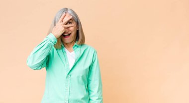 Orta yaşlı havalı bir kadın şok olmuş, korkmuş ya da korkmuş görünüyor. Yüzü elle kaplanmış ve parmaklarının arasından gizlice bakıyor.