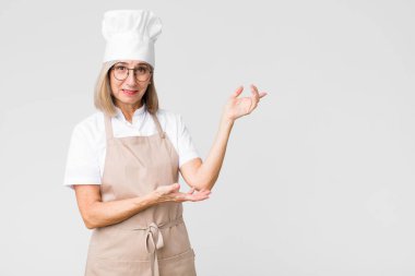 Orta yaşlı fırıncı kadın gururla ve güvenle gülümsüyor, mutlu ve tatmin olmuş hissediyor ve düz duvara karşı fotokopi alanında bir konsept gösteriyor.