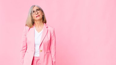 Endişeli, kafası karışık, budala bir ifadeyle orta yaşlı, havalı bir kadın kopya alanı arıyor, düz duvara karşı şüpheyle bakıyor.