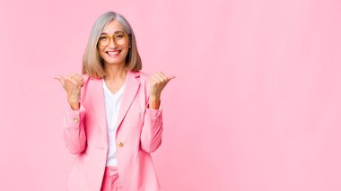 Orta yaşlı hoş bir kadın neşeli ve mutlu görünüyor, kaygısız ve pozitif hissediyor iki başparmağı düz duvara dayalı