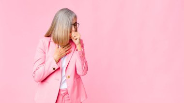 Orta yaşlı havalı bir kadın boğazı ağrıyan ve grip belirtileri gösteren, ağzı düz duvara dayalı öksüren hasta bir kadın.