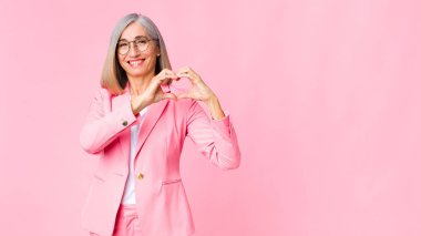 Orta yaşlı, havalı bir kadın gülümsüyor ve mutlu, sevimli, romantik ve aşık oluyor. İki elini de duvara dayayıp kalp şekli veriyor.