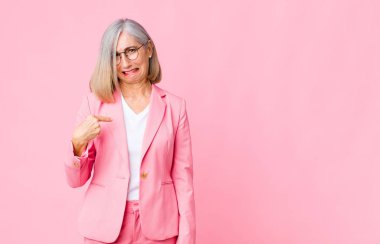 Orta yaşlı havalı bir kadın gururlu, kendinden emin ve mutlu görünüyor, gülümsüyor ve kendini işaret ediyor ya da düz duvara karşı bir numara işareti yapıyor.