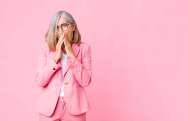 Orta yaşlı, havalı bir kadın endişeli, üzgün ve korkmuş, elleriyle ağzını kapatmış, endişeli görünüyor ve düz duvara çarpmış.