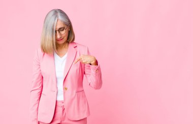 Orta yaşlı havalı bir kadın neşeli ve sıradan bir şekilde gülümsüyor, aşağıya bakıyor ve düz duvara doğru göğsünü gösteriyor.