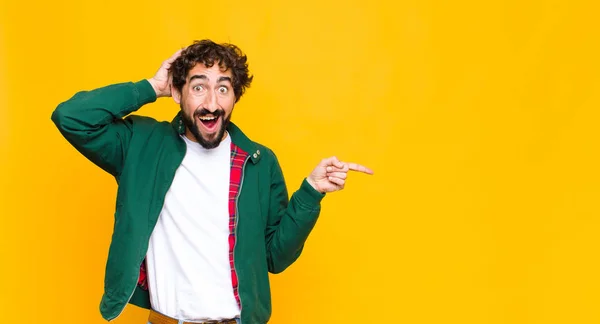 young crazy bearded man laughing, looking happy, positive and surprised, realizing a great idea pointing to lateral copy space against flat wall
