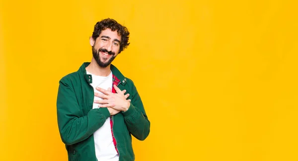 young crazy bearded man feeling romantic, happy and in love, smiling cheerfully and holding hands close to heart against flat wall