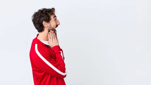 young bearded man back view feeling happy, excited and surprised, looking to the side with both hands on face against copy space wall