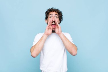 Genç çılgın sakallı adam mutlu, heyecanlı ve pozitif hissediyor. Elleri ağızda, düz duvara karşı bağırıyor.