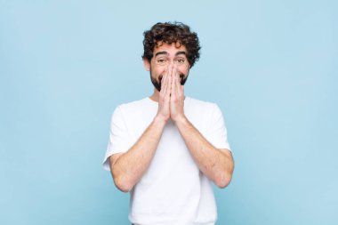 Genç çılgın sakallı adam mutlu ve heyecanlı, şaşırmış ve hayrete düşmüş elleriyle ağzını kapatmış, düz duvara karşı sevimli bir ifadeyle kıkırıyor.