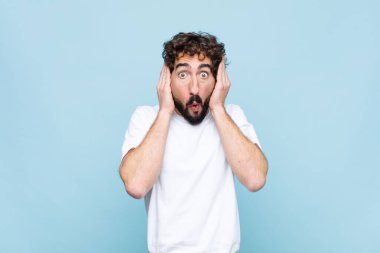 Genç çılgın sakallı adam hoş olmayan bir şekilde şok olmuş, korkmuş ya da endişeli görünüyor, ağzı sonuna kadar açık ve iki kulağını da düz duvara karşı kapatıyor.