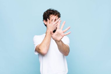 Genç çılgın sakallı adam yüzünü eliyle kapatıyor ve diğer elini kamerayı durdurmak için öne koyuyor, düz duvara karşı fotoğraf veya resimleri reddediyor.